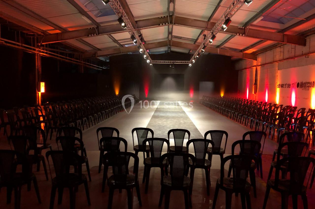 Salle industrielle aménagée avec une allée centrale éclairée, entourée de rangées de chaises noires en plastique.