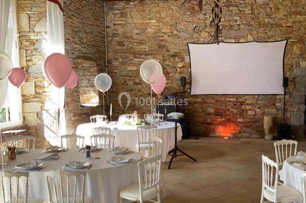 Salle en pierre décorée pour un événement, avec tables dressées, ballons roses et blancs, et un écran de projection.