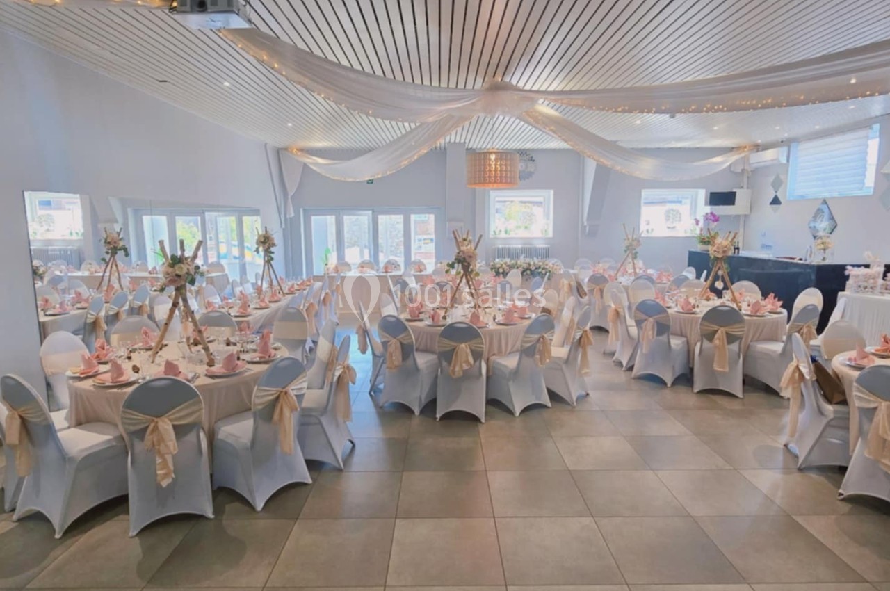 Salle de réception décorée avec des tables rondes, nappes blanches, chaises ornées de nœuds et éclairage chaleureux.