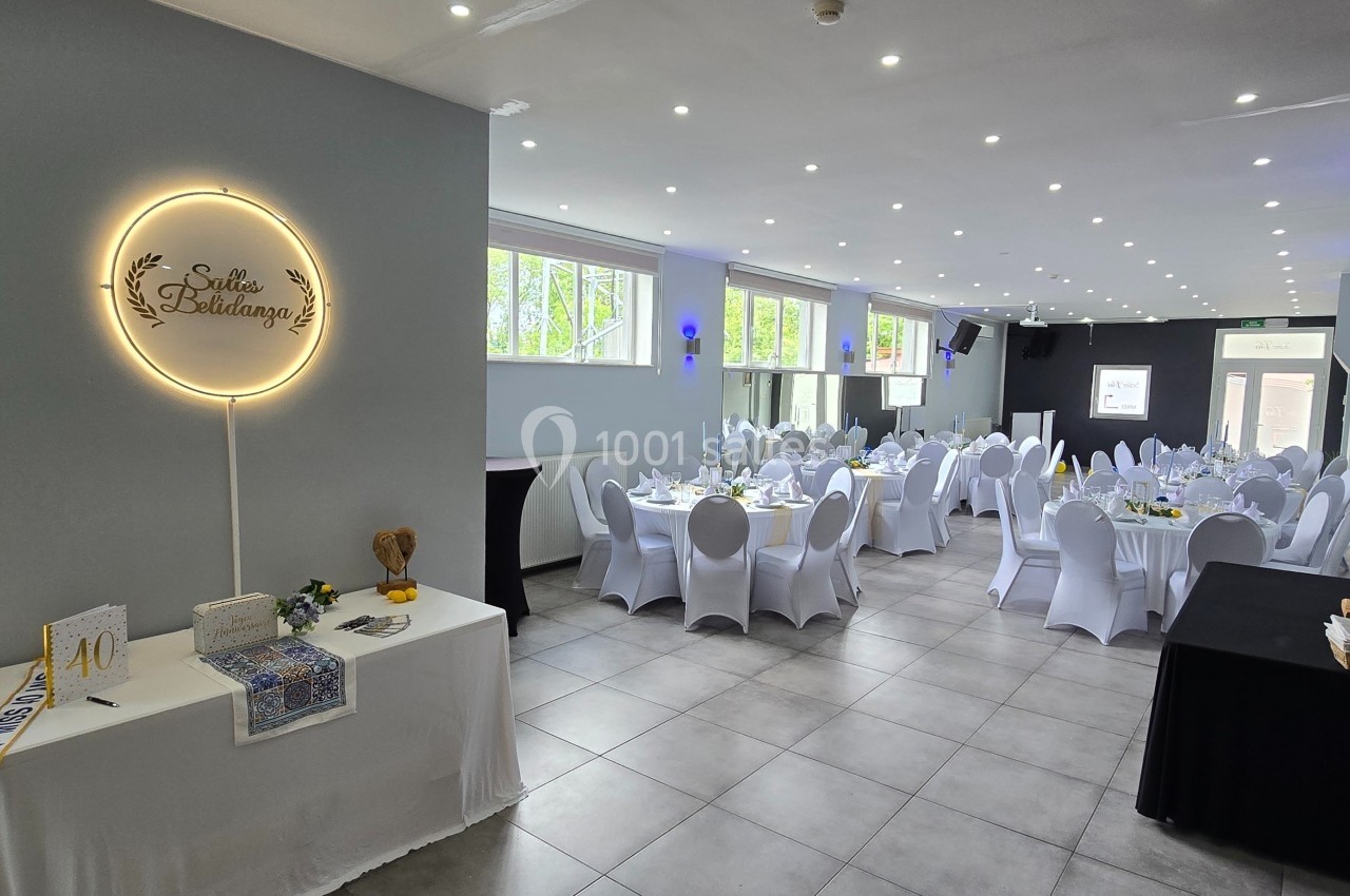 Salle de réception décorée avec des tables rondes dressées, chaises blanches et éclairage lumineux.