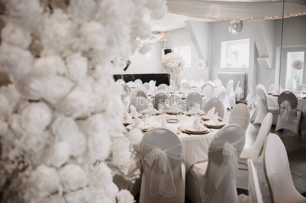 Salle de réception décorée pour un mariage avec des tables rondes, nappes blanches et chaises ornées de nœuds en organza.