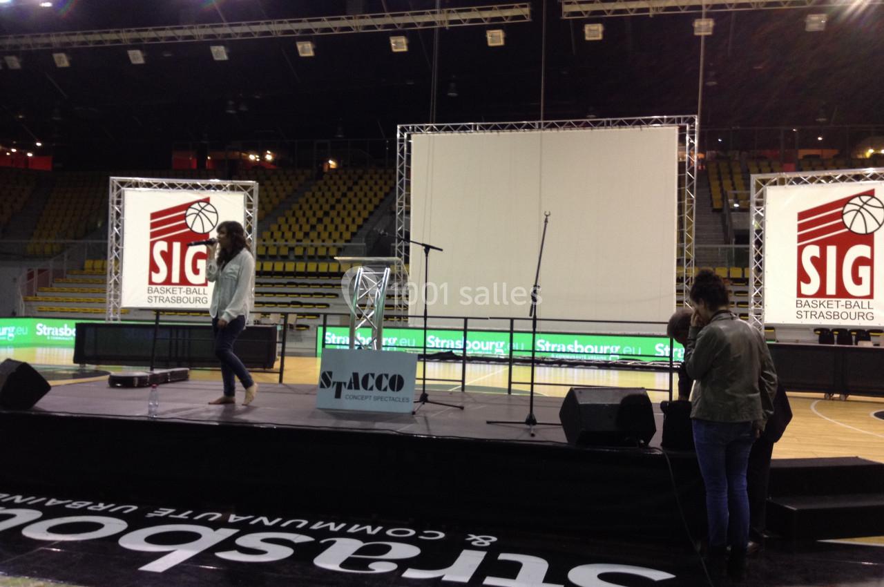 Scène avec micros et écran géant dans un stade de basket, préparatifs en cours avec deux personnes sur le plateau.