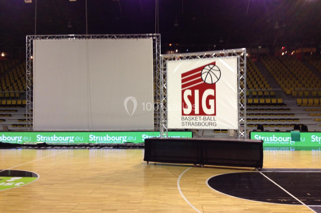 Écran géant et panneau SIG Basket-Ball Strasbourg installés sur un terrain de basket dans une salle vide.