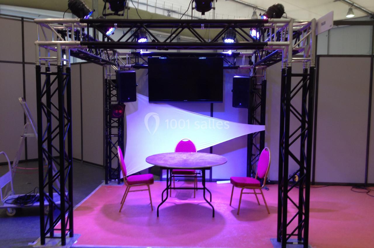 Structure métallique avec éclairage, deux chaises roses et une table ronde sur un tapis rose dans un espace intérieur.