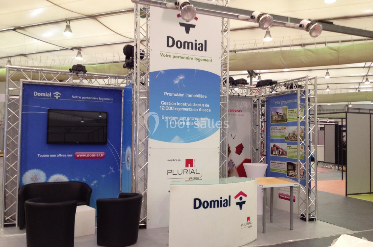 Stand d'exposition avec affiches promotionnelles, mobilier et écran, représentant une entreprise immobilière.