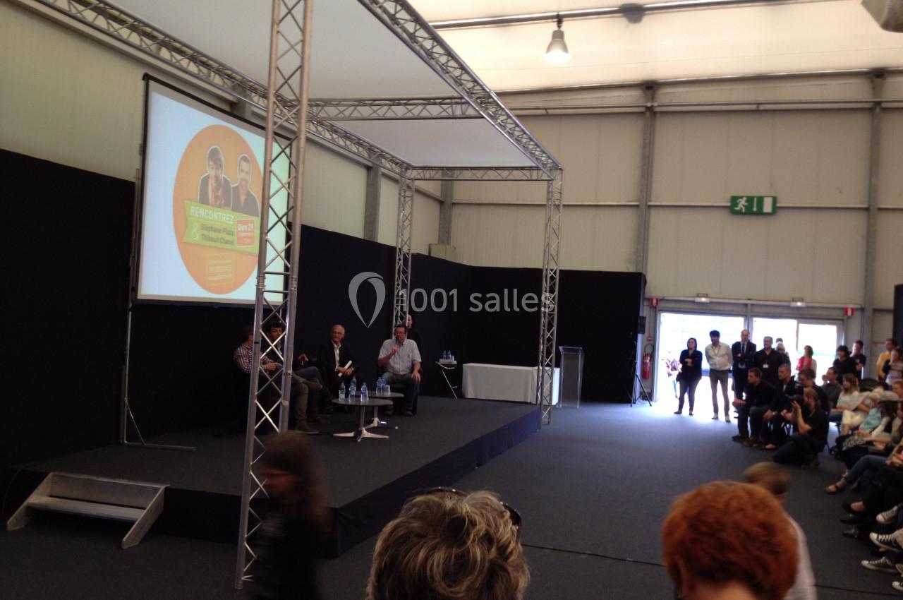 Un panel de trois intervenants discute sur une scène dans une salle avec un public assis et debout.