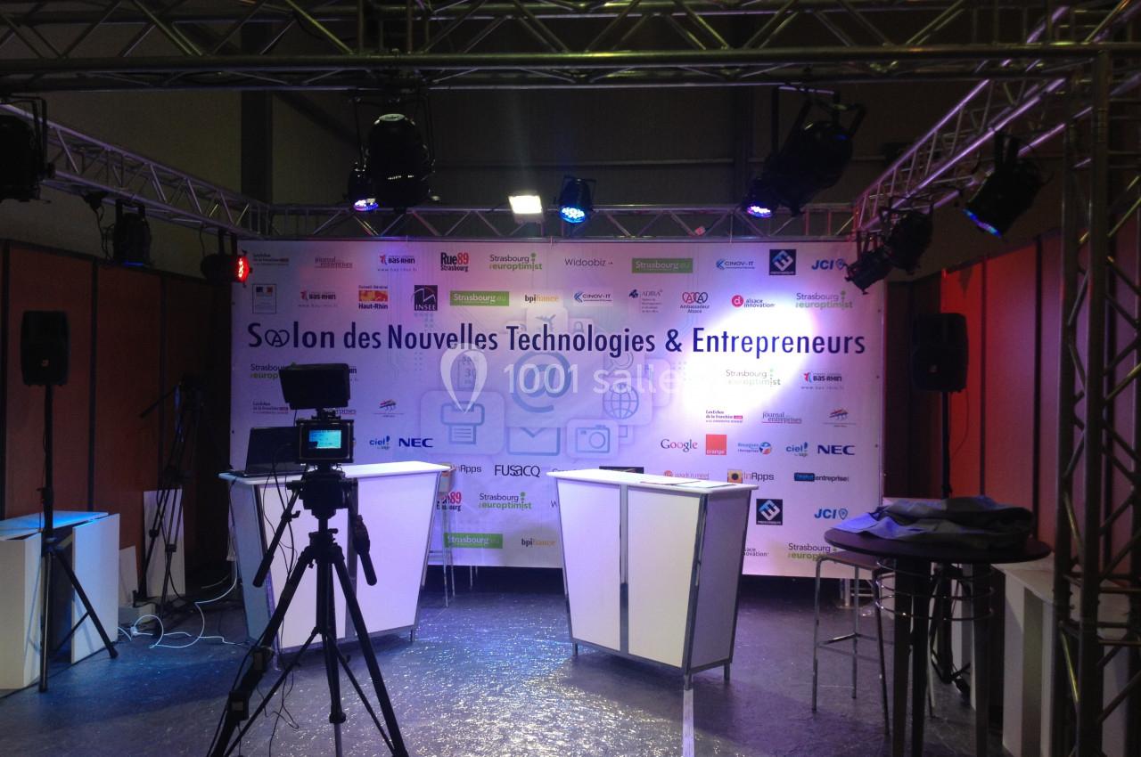 Scène d'un salon professionnel avec un fond promotionnel, des projecteurs et une caméra sur trépied.