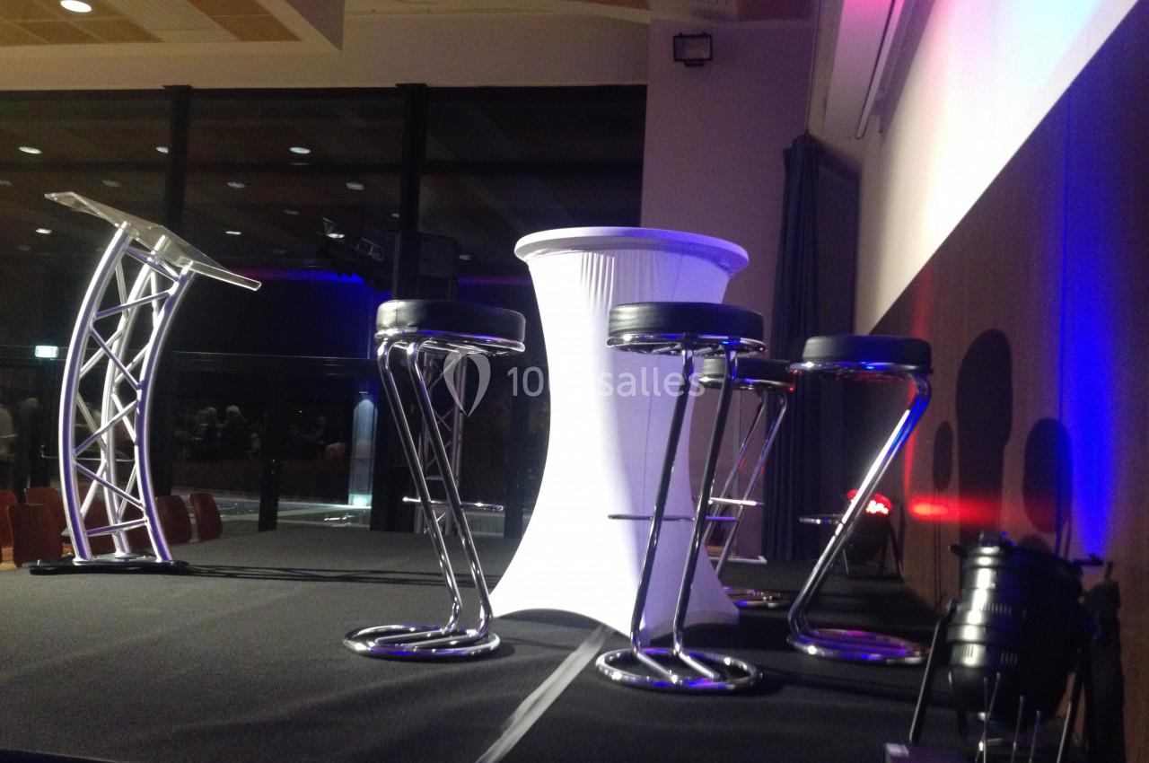 Scène avec pupitre, table haute blanche et tabourets noirs, éclairée par des projecteurs dans une salle vitrée.