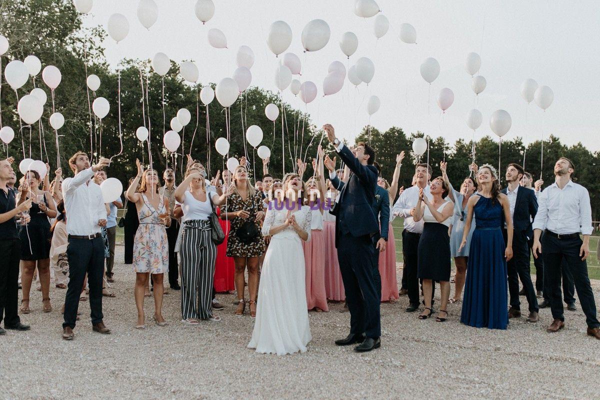 Un groupe de personnes en tenue de cérémonie lâche des ballons blancs et roses en extérieur.
