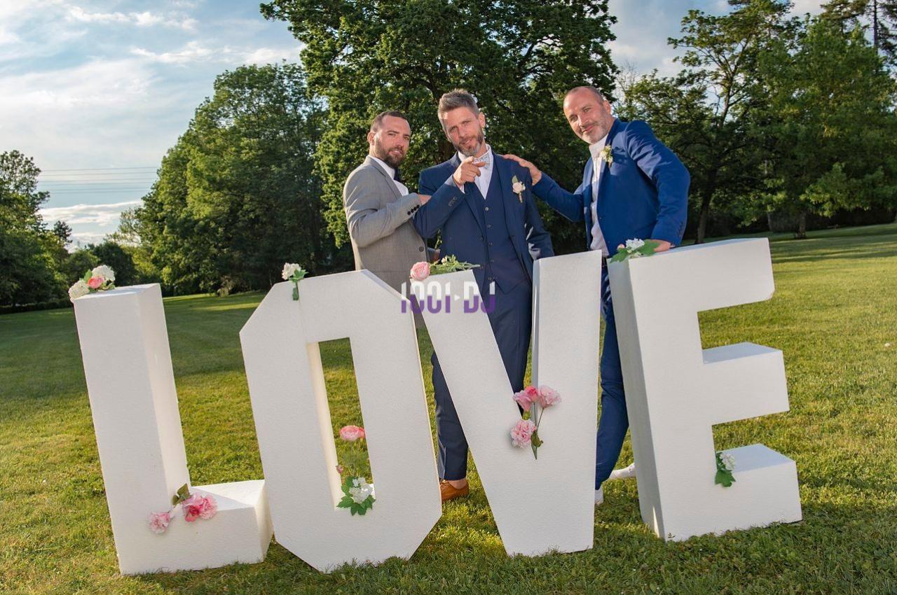 Trois hommes en costume posent devant de grandes lettres blanches formant le mot ’LOVE’ dans un parc verdoyant.