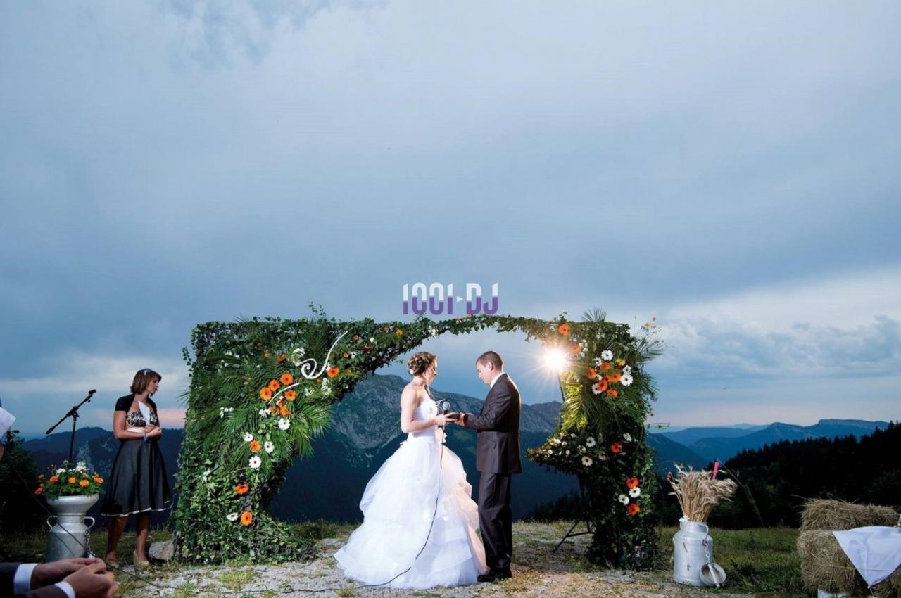 Un couple échange ses vœux sous une arche fleurie en extérieur, avec une vue sur des montagnes en arrière-plan.