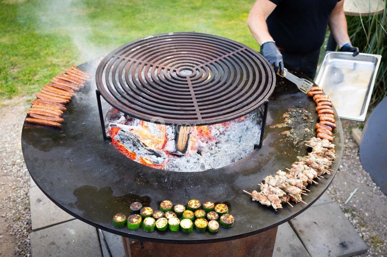 Grill circulaire en plein air avec saucisses, brochettes et légumes cuisant sur une plaque chauffée au feu de bois.