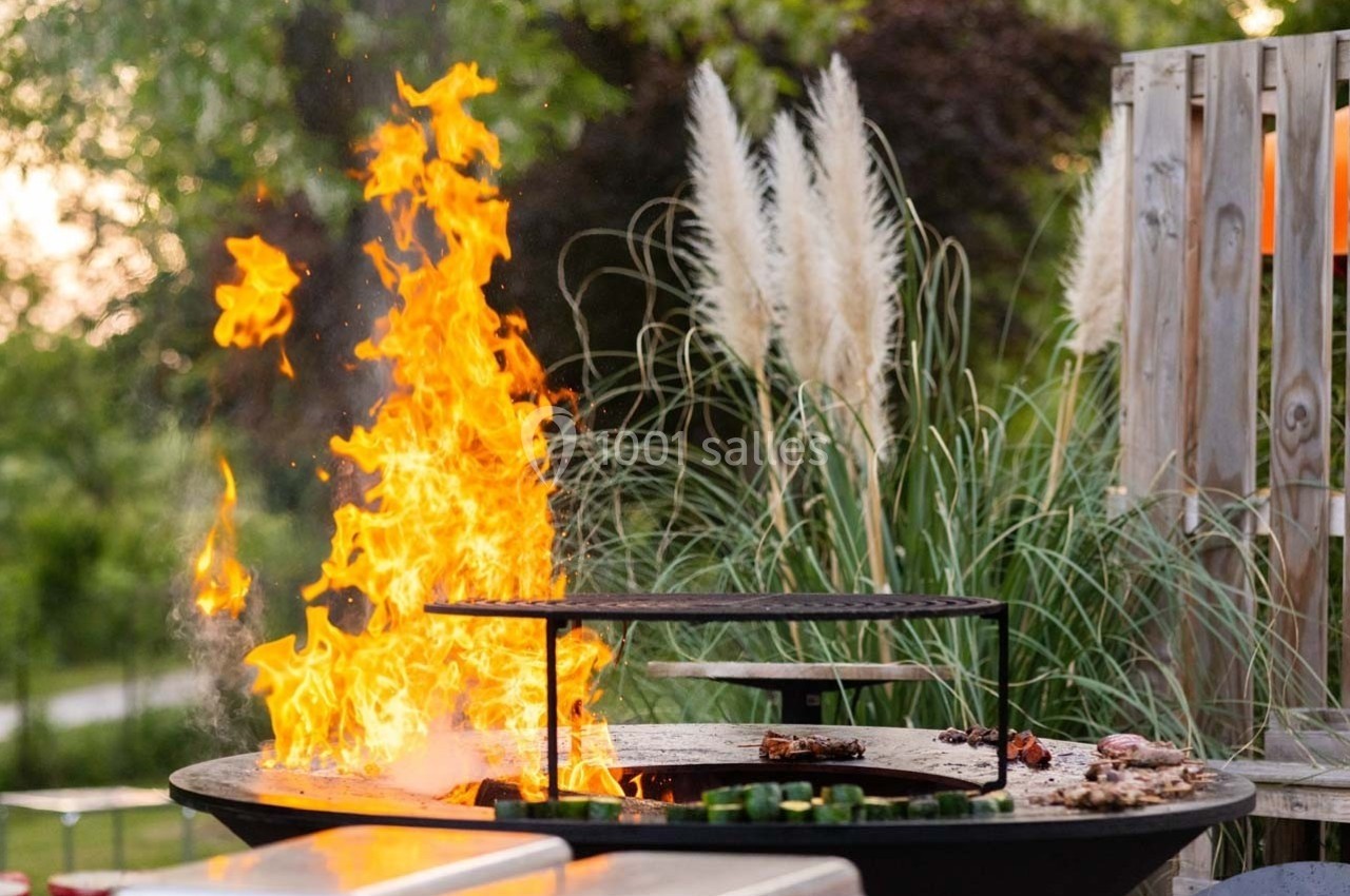 Barbecue circulaire en plein air avec des flammes vives, entouré de végétation et de mobilier de jardin.
