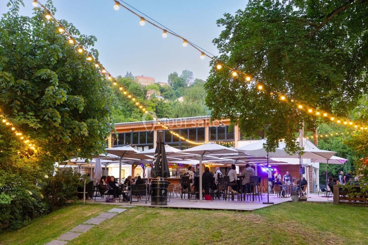 Terrasse extérieure d'un restaurant avec des guirlandes lumineuses, des tables occupées et entourée de verdure.