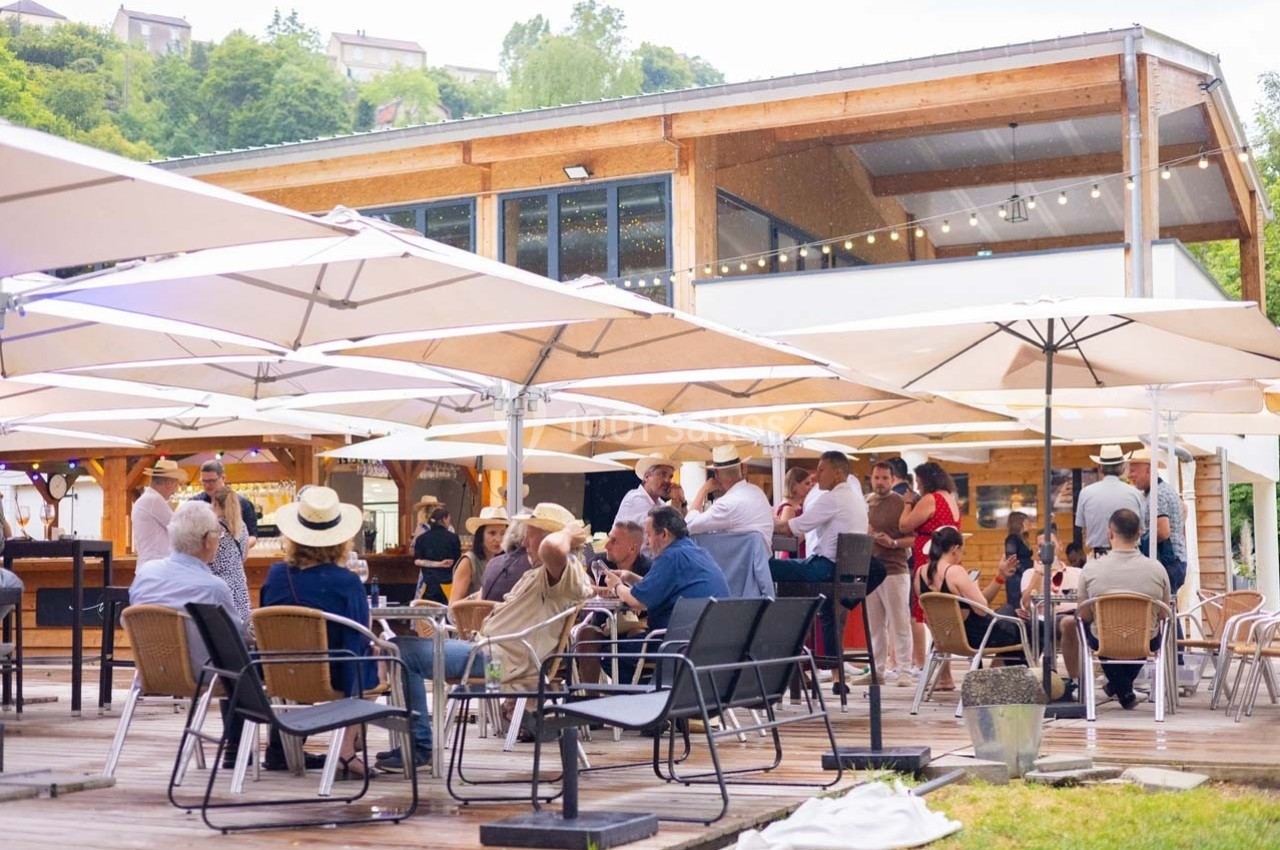 Personnes assises et debout sur une terrasse en bois, protégées par des parasols, devant un bâtiment moderne en bois.