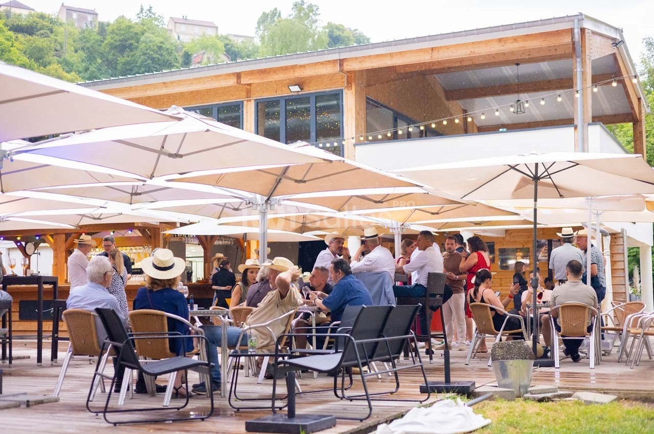 Personnes assises et debout sur une terrasse en bois, protégées par des parasols, devant un bâtiment moderne en bois.
