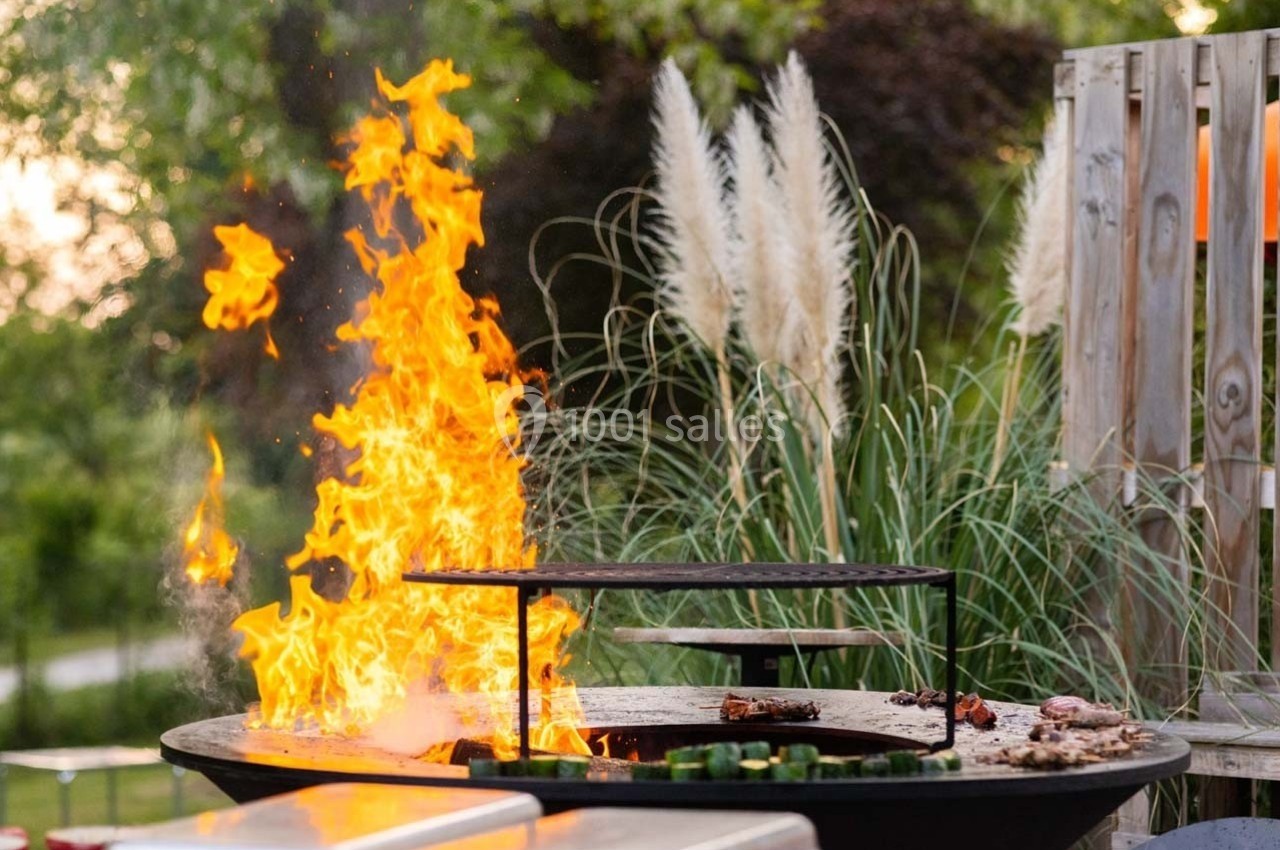 Flammes vives s'élevant d'un barbecue circulaire en plein air, entouré de végétation et de décorations naturelles.