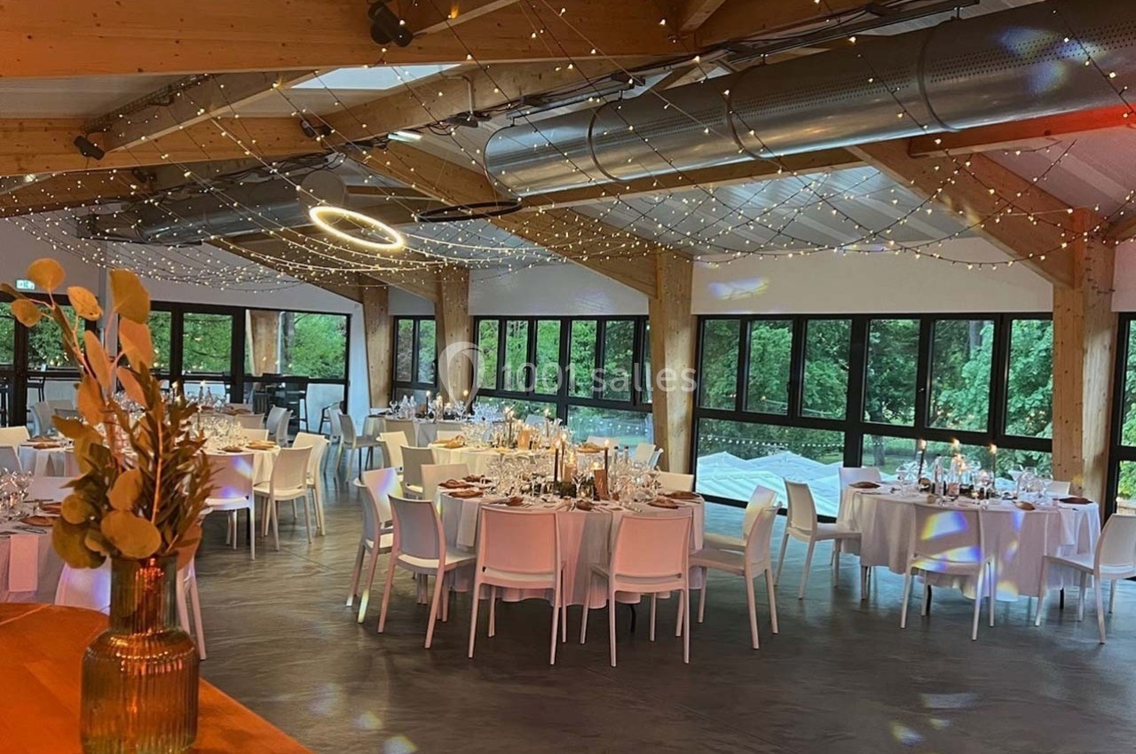 Salle de réception lumineuse avec tables dressées, grandes baies vitrées et guirlandes lumineuses suspendues.