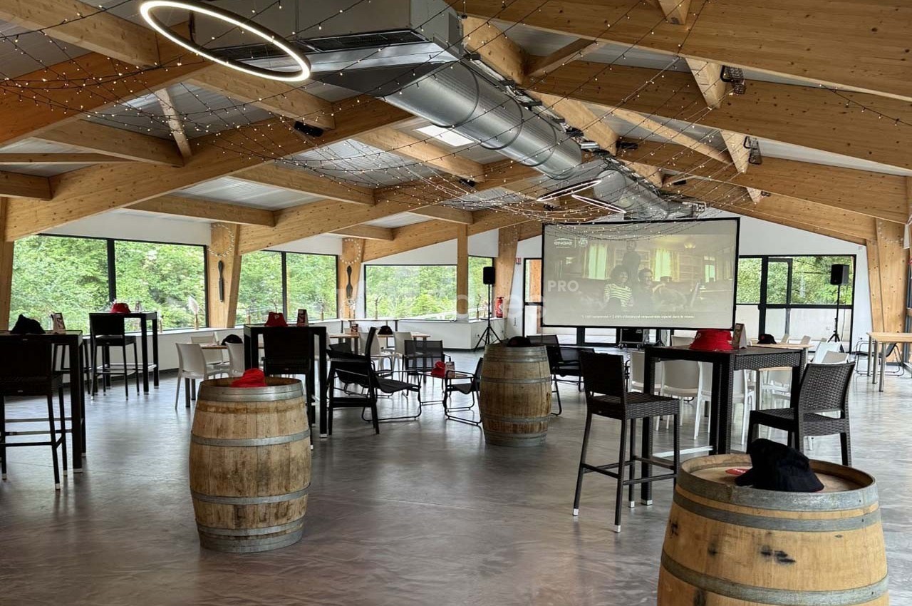 Salle lumineuse avec charpente en bois, barils en décoration, tables hautes et écran de projection allumé.