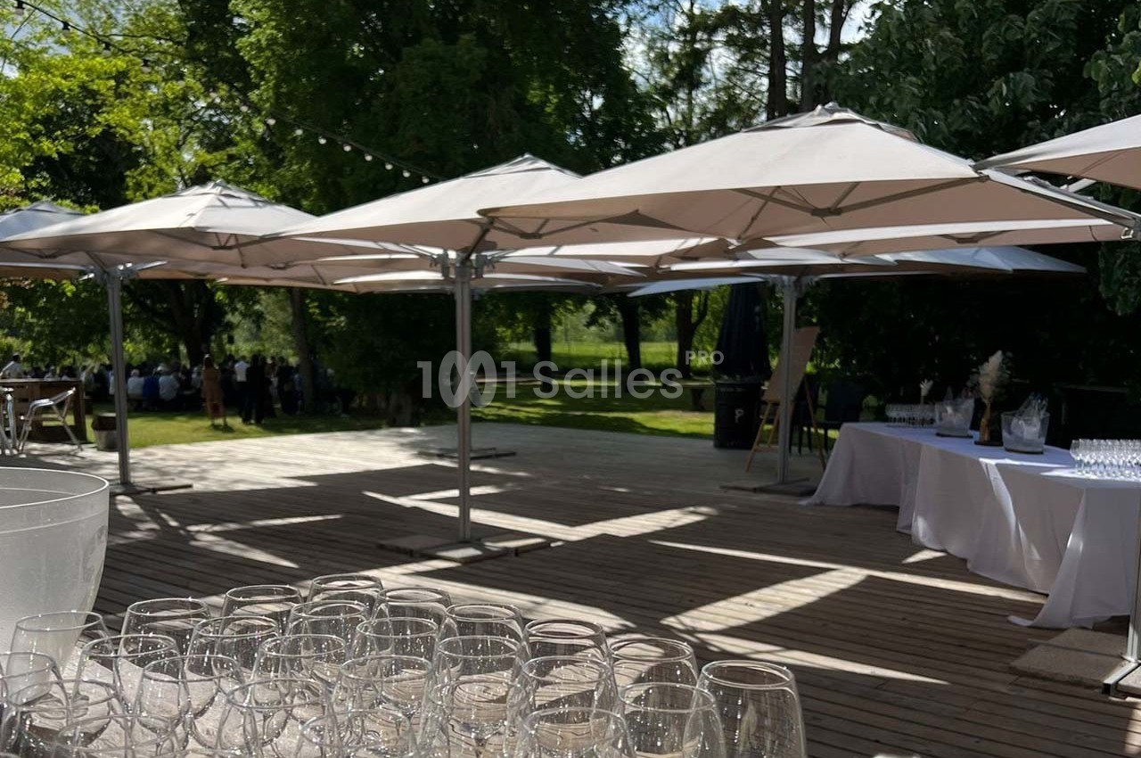 Terrasse en bois avec des parasols blancs, entourée de verdure, tables dressées et verres alignés au premier plan.