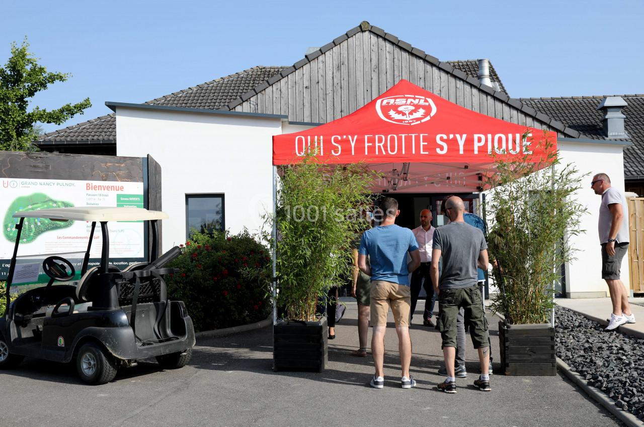Entrée d'un bâtiment avec une tente rouge, des plantes en pot et des personnes discutant à proximité d'une voiturette de…