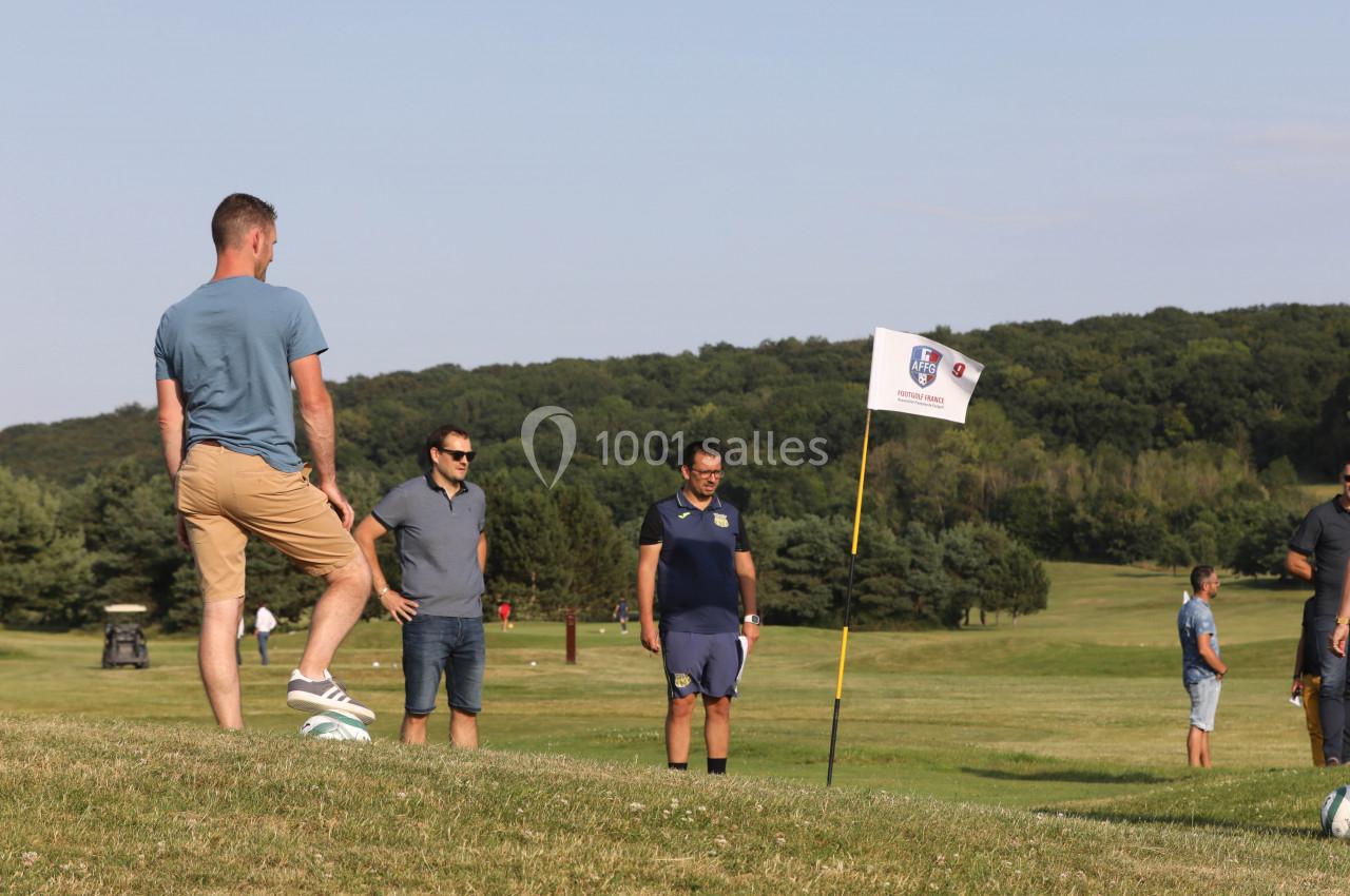 Un homme joue au football sur un terrain herbeux, observé par trois personnes près d'un drapeau.