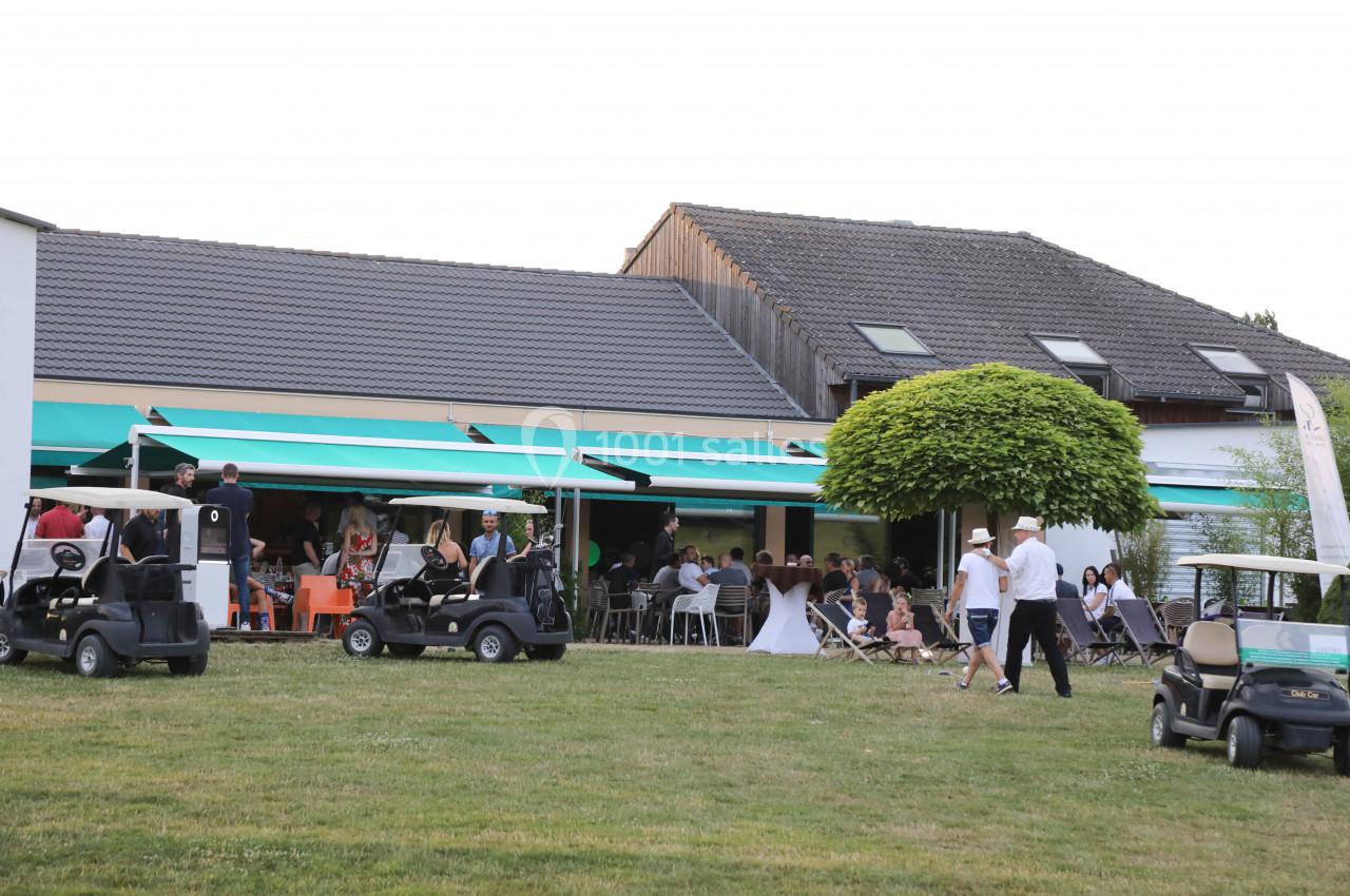 Terrasse d'un bâtiment avec des tables occupées, des personnes debout et des voiturettes de golf stationnées sur la pelouse.