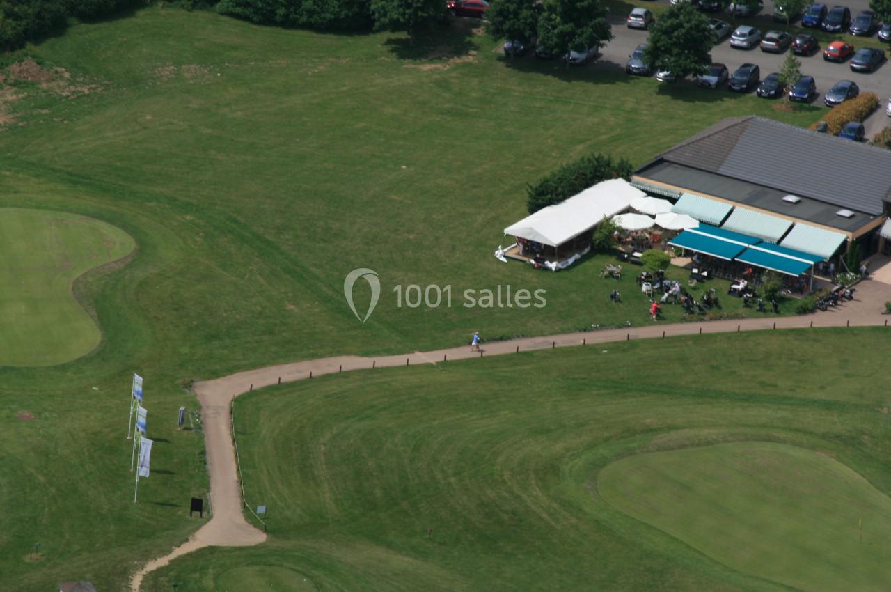 Vue aérienne d'un terrain de golf avec un bâtiment, une terrasse couverte et des voitures garées à proximité.
