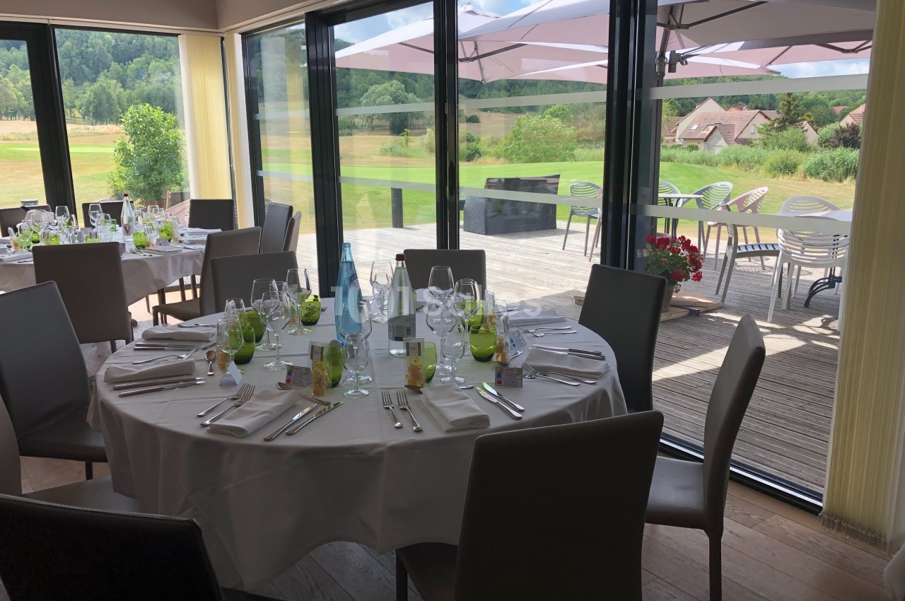 Salle de restaurant lumineuse avec tables dressées, vue sur une terrasse et un paysage verdoyant à travers de grandes baies…