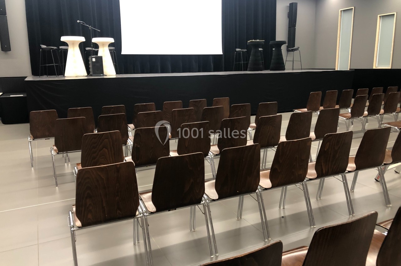 Salle de conférence avec des rangées de chaises en bois face à une scène équipée de micros et de tabourets.