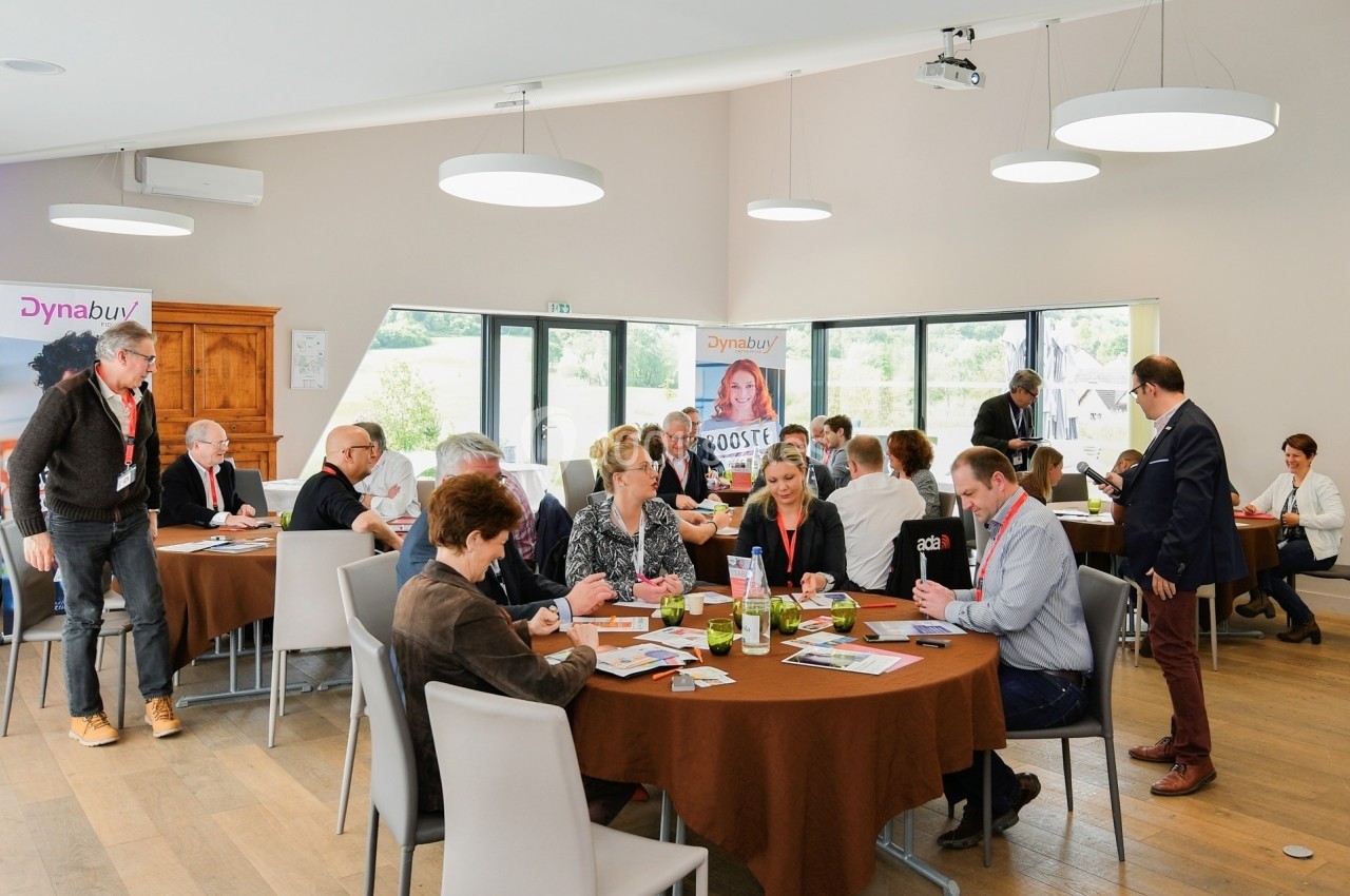 Personnes échangeant autour de tables lors d'un événement professionnel dans une salle lumineuse.
