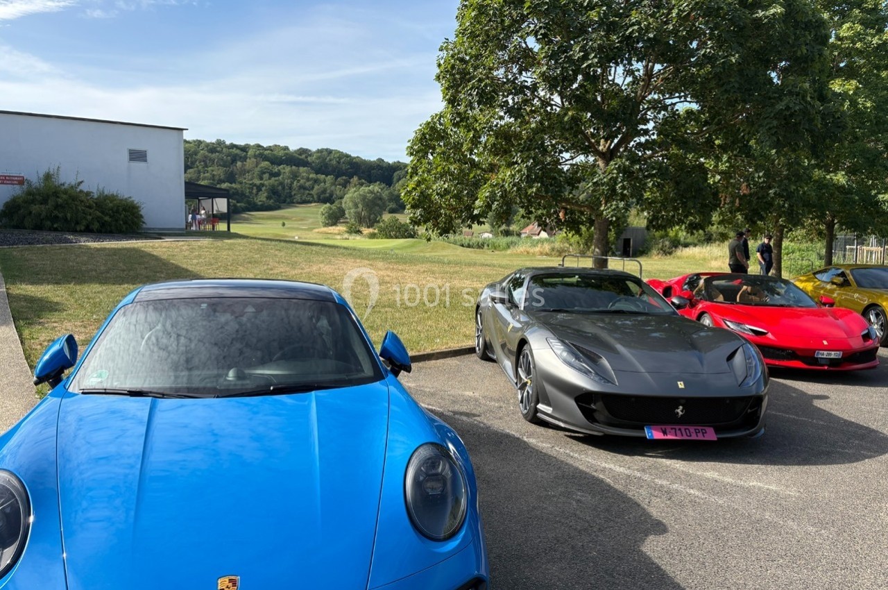 Trois voitures de sport, une Porsche bleue et deux Ferrari grises et rouges, stationnées sur un parking en plein air.
