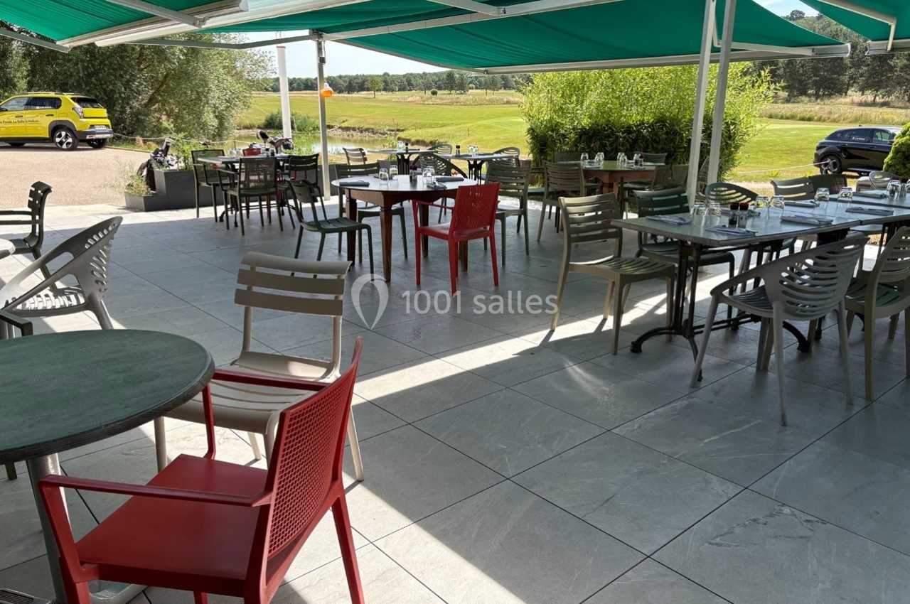 Terrasse extérieure avec tables et chaises disposées sous des stores verts, vue sur un paysage champêtre.