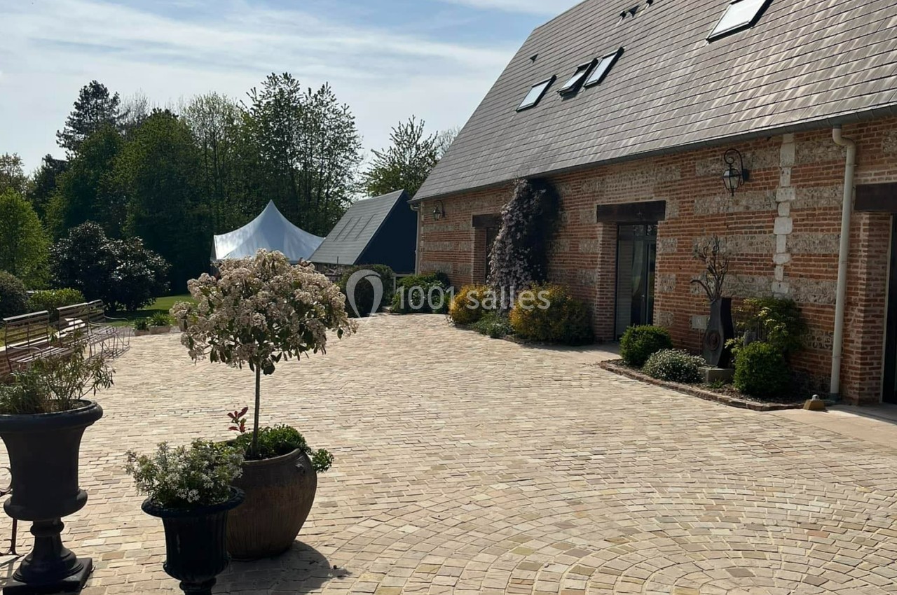 Cour pavée devant une maison en briques avec toit en ardoise, entourée de végétation et de pots de fleurs.