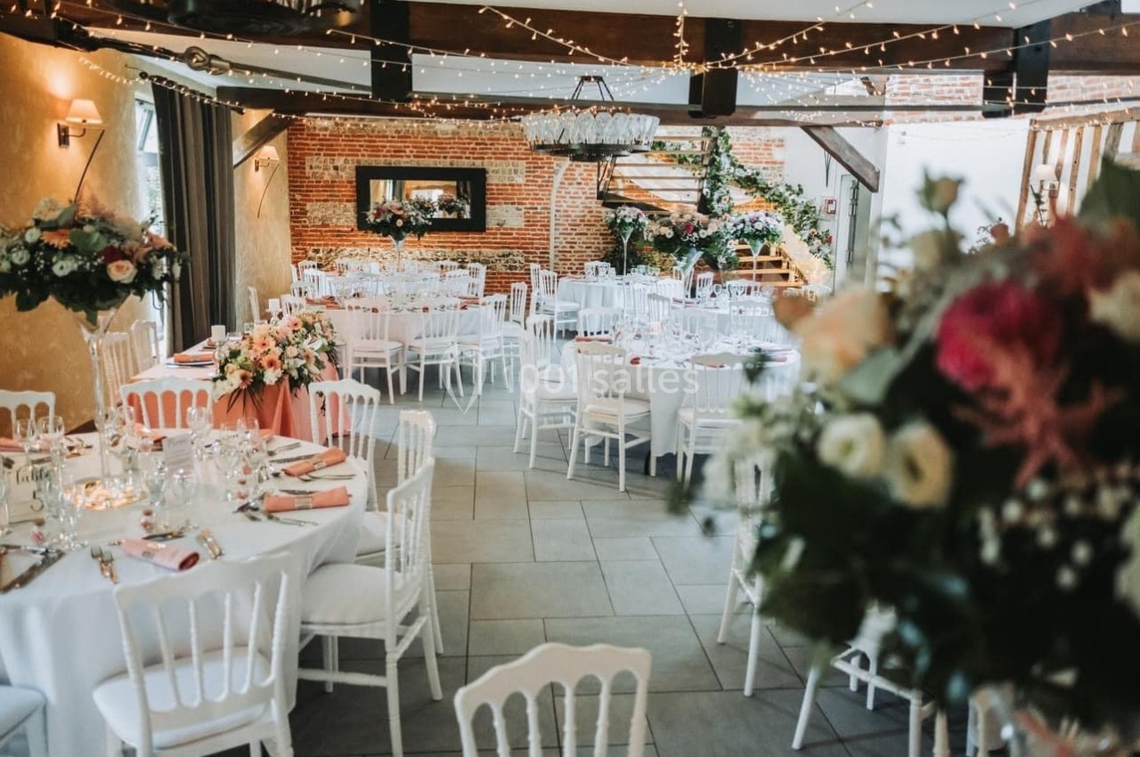 Salle de réception décorée avec des tables rondes, nappes blanches, fleurs et guirlandes lumineuses suspendues.