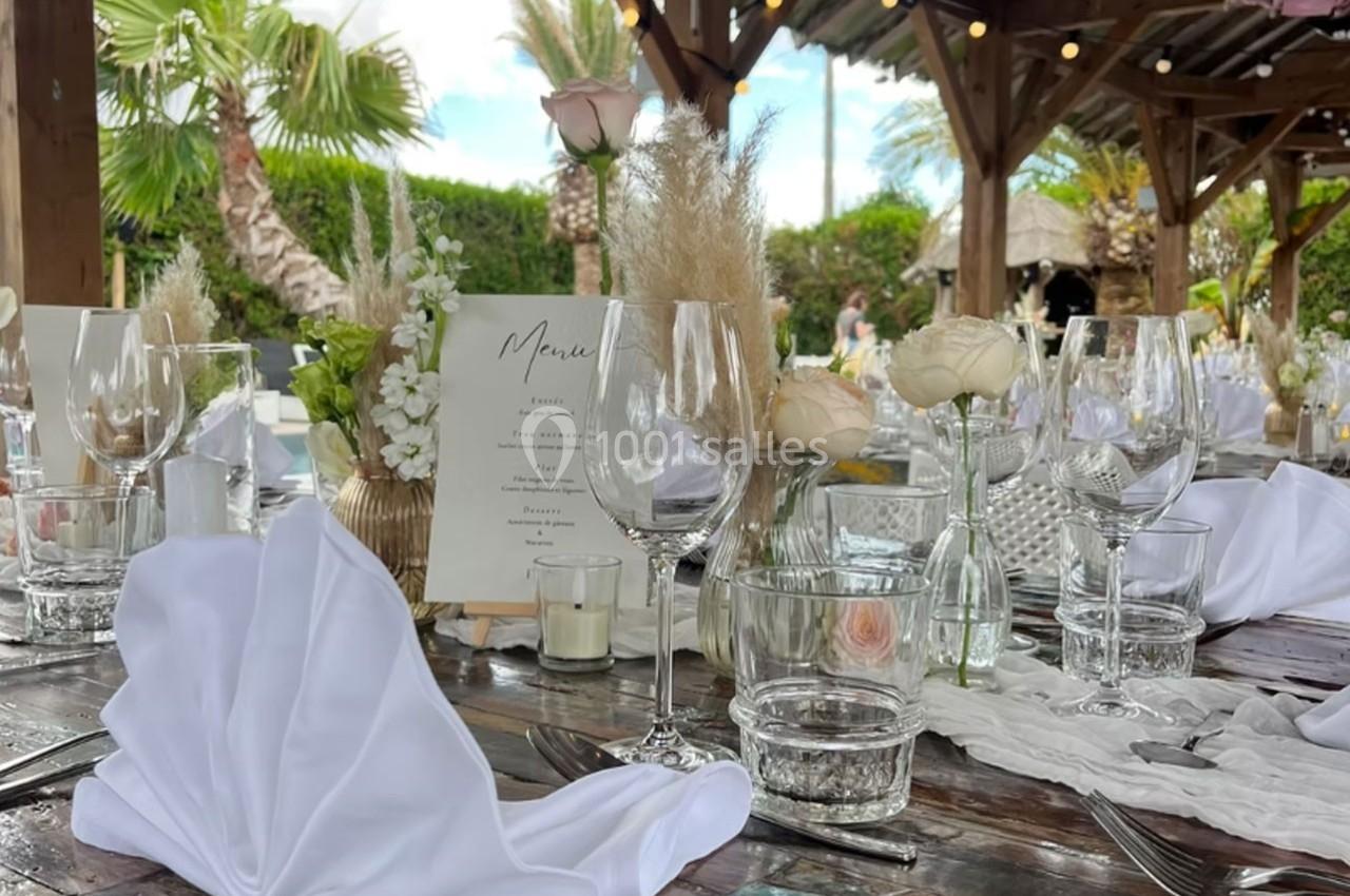 Table dressée pour un repas en extérieur avec nappes blanches, verres, fleurs et menu sous un abri en bois.