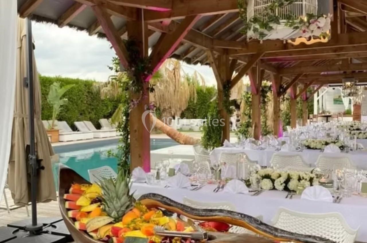 Salle de réception en plein air avec tables dressées, décoration florale et buffet de fruits près d'une piscine.