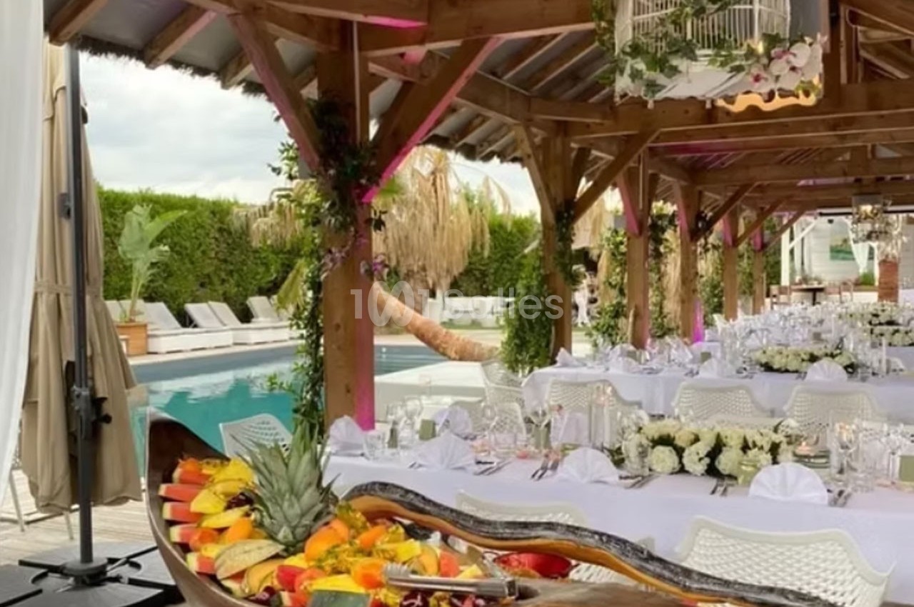 Salle de réception en plein air avec tables dressées, décoration florale et buffet de fruits près d'une piscine.