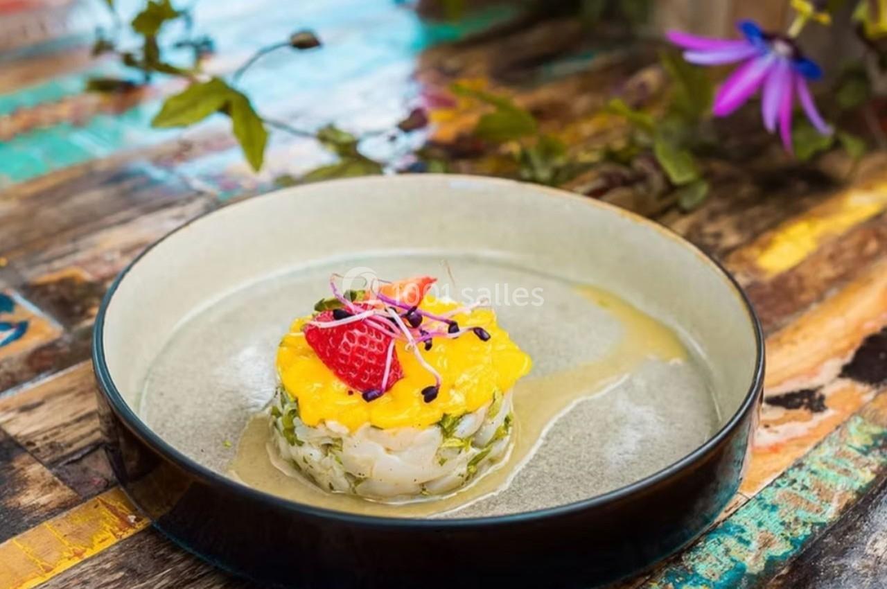 Tartare de poisson garni de mangue, fraises et pousses, présenté dans une assiette sur une table en bois colorée.