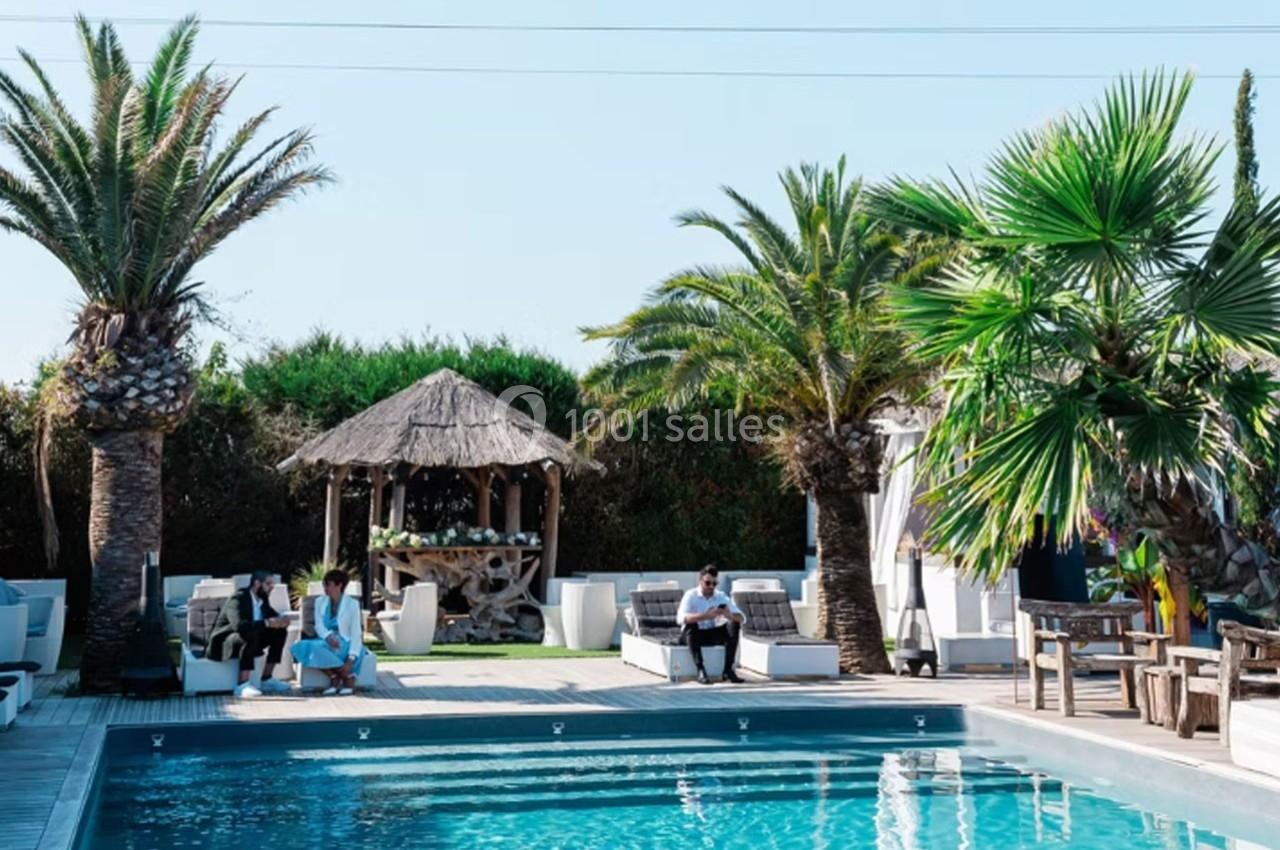 Piscine extérieure entourée de palmiers, avec des chaises longues et un kiosque en bois au fond.