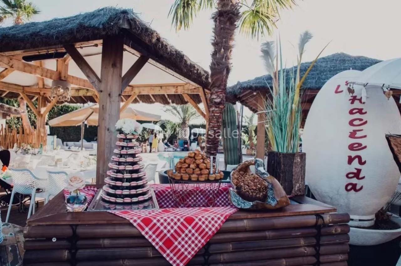 Stand extérieur avec présentation de desserts, pain et viennoiseries sur une table décorée, entouré de paillotes.