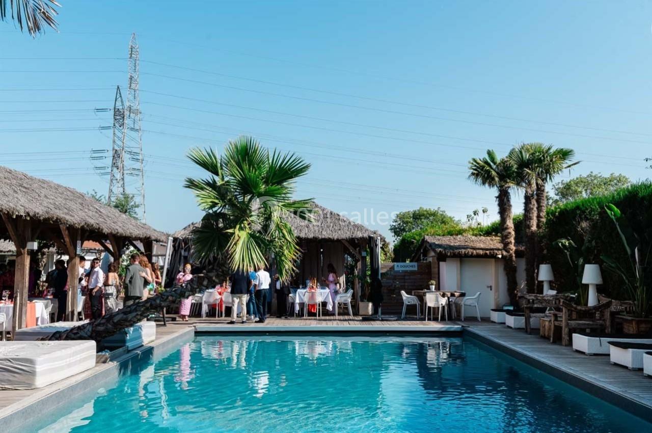 Piscine extérieure entourée de terrasses en bois, de palmiers et de cabanes avec des personnes assises et debout.