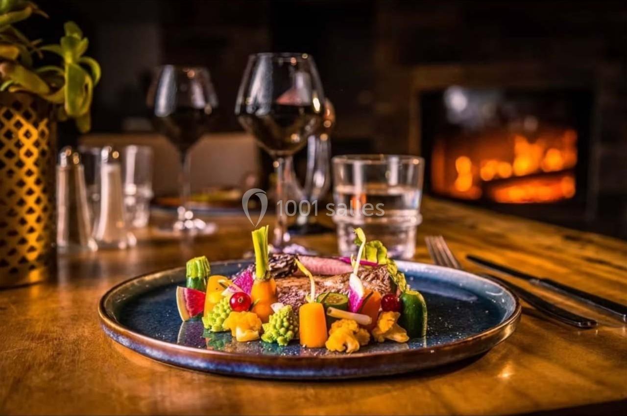 Assiette gastronomique colorée avec légumes et poisson, servie sur une table en bois près d'un feu de cheminée.