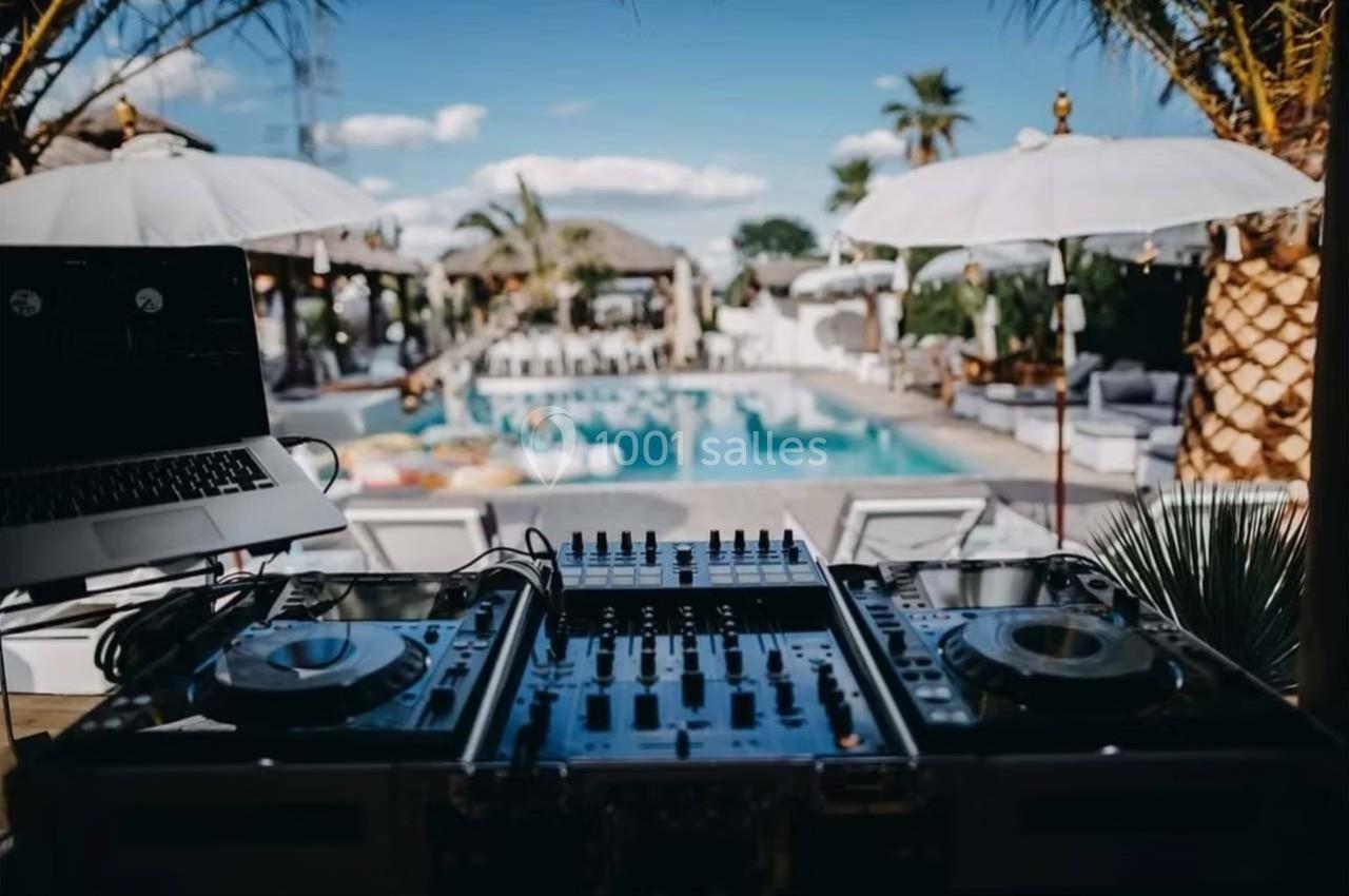 Table de mixage avec platines et ordinateur portable en premier plan, vue sur une piscine entourée de parasols et palmiers.