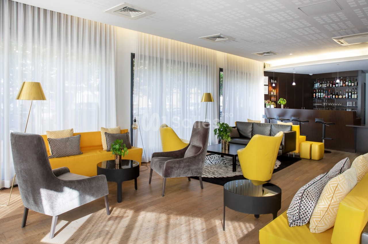 Salon moderne avec fauteuils jaunes et gris, tables basses noires, grandes fenêtres avec rideaux blancs et un bar en arrière…