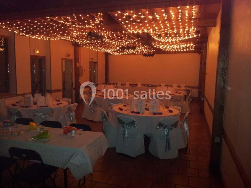 Salle de réception décorée avec des guirlandes lumineuses au plafond et des tables rondes dressées pour un événement.