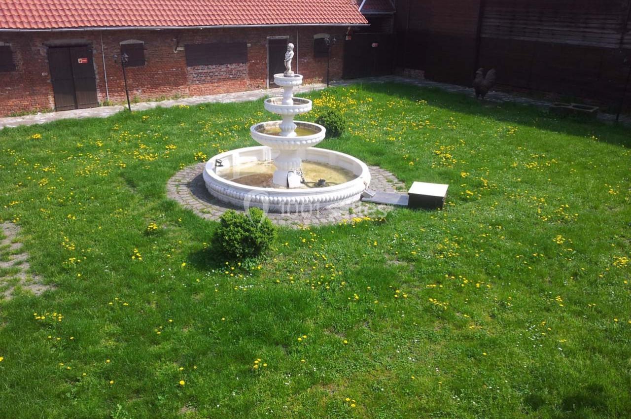 Fontaine blanche à trois niveaux entourée d'une pelouse verte parsemée de pissenlits, dans une cour bordée de bâtiments en…