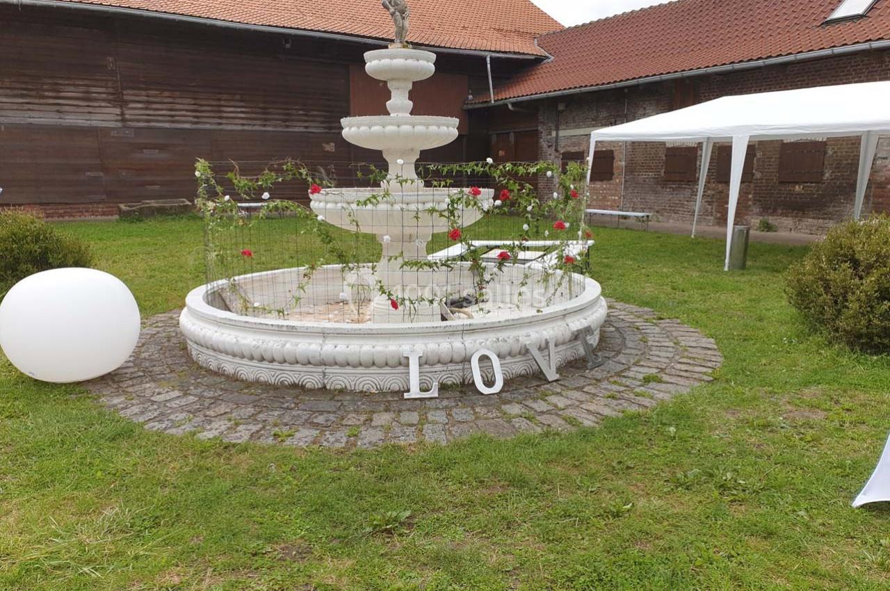 Fontaine blanche décorée de roses rouges, entourée de pelouse, avec des lettres formant ’LOVE’ au sol.