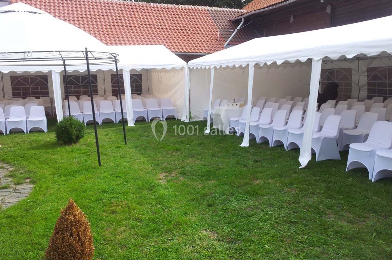 Chaises blanches disposées sous des tentes dans une cour avec pelouse, entourée de bâtiments en briques et bois.