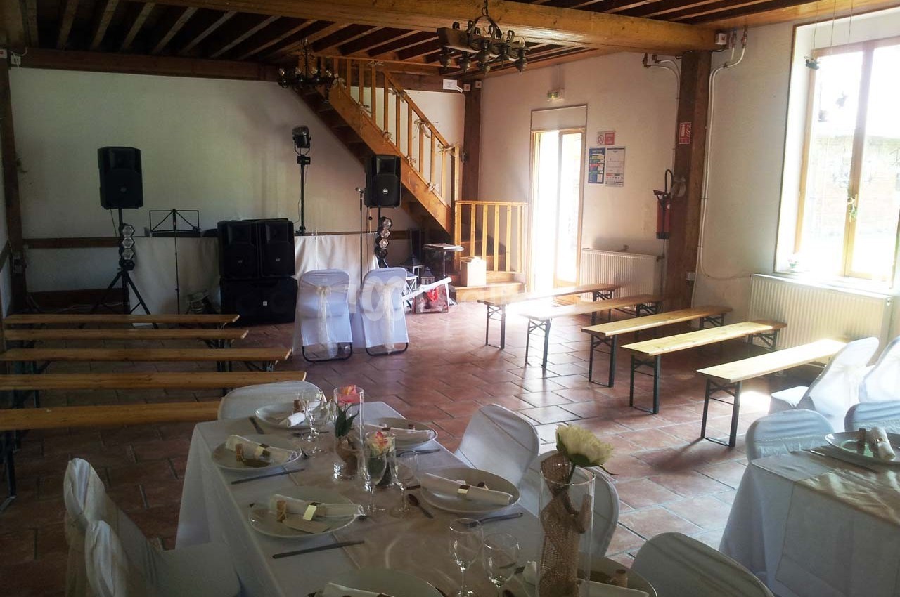 Salle de réception avec tables dressées, chaises blanches, bancs en bois et équipement audio près d'un escalier en bois.